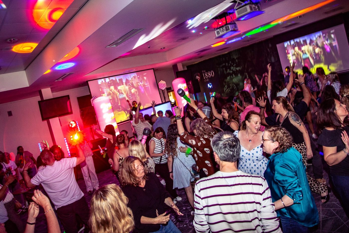 A packed dancefloor with arms raised and music videos on the big screens